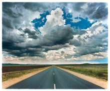 Load image into Gallery viewer, Photography by Richard Heeps. A photograph looking along a long straight road into the distance, it is edged either side with sand and then prairie land. The vast, cloudy sky sits above.