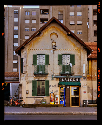 Photograph by Richard Heeps. A traditional Italian Tobacconist shop, here in a Swiss Cottage style building set against a vast urban apartment block.