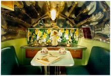 Load image into Gallery viewer, Photograph by Richard Heeps. Inside a trailer, there is a fixed table and chairs with two tea cups and saucers on the table. Behind on the shelf is a tea set and a doll dressed as a bride.