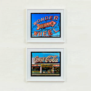 Two framed vintage-style signs on a white wall, one with 'Wonder Wheel' and the other with 'Coca-Cola' artworks by Richard Heeps