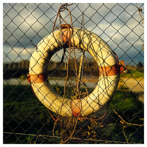 Photograph by Richard Heeps.  A lifebuoy sits hanging behind a wire fence. Water and a cloudy sky are blurred behind.