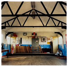 Load image into Gallery viewer, Photograph by Richard Heeps.  Inside a Scout Hut, cabinets for Beavers, Cubs and Scouts sit on a slightly raised platform along with wooden folding chairs.  There are dark wooden beams contrasting against the white ceiling.
