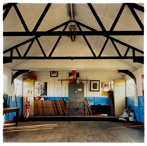 Photograph by Richard Heeps.  Inside a Scout Hut, cabinets for Beavers, Cubs and Scouts sit on a slightly raised platform along with wooden folding chairs.  There are dark wooden beams contrasting against the white ceiling.