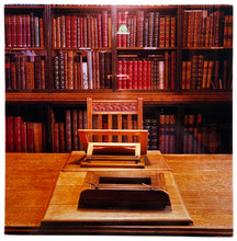 Load image into Gallery viewer, Photograph by Richard Heeps.  In the library with the books on the shelf behind and in foreground a wooden table with a chair.  The table has two easels lined up with the chair, the easel nearest the chair is open.