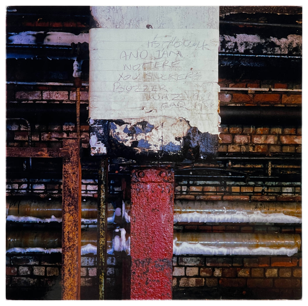 Photograph by Richard Heeps. Warm-toned bricks and metal pipes with a white taped red downpipe at the photo's centre.  Handwriting on the white tape reads Buzzer Was Ear.