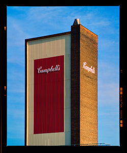 Photograph by Richard Heeps.  The Campbell's factory tower, cream and red with the Campbell's logo in the middle, against a blue sky.