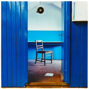 Photograph by Richard Heeps. A photograph of chair which sits in a derelict factory in a colour block blue painted room, the perspective creates depth.
