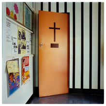 Load image into Gallery viewer, A photograph by Richard Heeps depicting a wooden door with a cross on it sitting slightly ajar set in a black and white striped wall. On the left hand side sits local community posters.