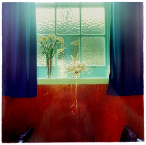 Photograph by Richard Heeps.  A bunch of white carnations in a glass vase together with another bunch of white flowers sit on a windowsill in front of a frosted glass window.  The curtains are by contrast deep blue and the wall is a burnt orange colour.  
