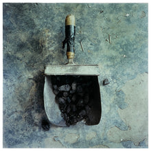 Load image into Gallery viewer, A photograph of a battered coal scuttle on the floor filled with coal, with its wooden handle taped. The coal scuttle sits on grey broken flooring. Photographer Richard Heeps