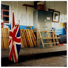 Load image into Gallery viewer, Photograph by Richard Heeps.  Inside a Cub Scout Hut, with the Union Jack Flag hanging and touching the floor.  The back half of the photo has a wooden platform with folding chairs on and the cubs own metal container.