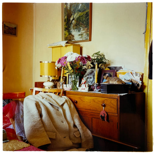 Photograph by Richard Heeps. A wooden dresser with photos, bunches of flowers, candles, ornaments, lamps and a full inbox on top.  In front of this busy piece of furniture are clothes laid on a chair, on top a white fur jacket. 