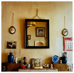 Photograph by Richard Heeps. A tiled mantelpiece full of paraphernalia from the past: clocks; porcelain animals and curios all seemingly randomly displayed.  There is a square mirror over the mantelpiece reflecting the room and on the far right of the photo is a red and white calendar, displaying July 1988. 