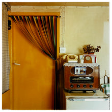 Load image into Gallery viewer, Photograph by Richard Heeps.  A vintage kitchen with an orange door and a plastic cream handle.  A door curtain is tied up over to the side of the door.  There sits to the right of the door a radio with a postcard from Largs tucked in the front.  On top of the radio is a doily on top of which sits a dial phone, carriage clock and houseplant sitting on top of the radio.