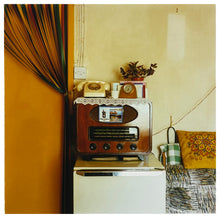 Load image into Gallery viewer, Photograph by Richard Heeps. This 1990s scene has a wooden-cased radio sitting on top of a fridge. The radio is used as a shelf for a dial telephone, a pot plant and various other items. There is a tied-up door curtain to the left and cushions to the right.