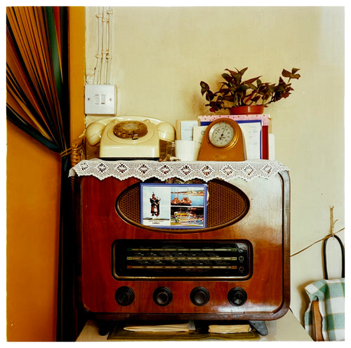 Photograph by Richard Heeps. Daisy's radio in its wooden casing sits in the middle of this photograph.  There is a postcard from Largs stuck on the radio speakers and on top of the radio the surface is crowded with houseplants, a phone, a mantel clock and other oddments. 