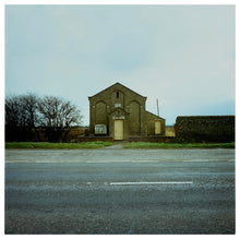 Load image into Gallery viewer, Photograph by Richard Heeps.  In the foreground is an empty road, on the opposite side of the road is a grass verge and then a yellow-doored, windowless, Chapel sitting solitary with no other buildings nearby.