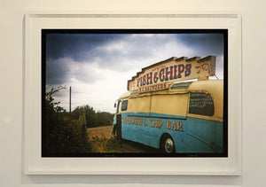 A moody Fenland sky is backdrop to a classic British Fish & Chips Van. The typography and the van itself has a fun fairground feel, contrary to the muted tones of this piece. Part of Richard Heeps series, 'A View of the Fens from the Car with Wings'.