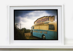 A moody Fenland sky is backdrop to a classic British Fish & Chips Van. The typography and the van itself has a fun fairground feel, contrary to the muted tones of this piece. Part of Richard Heeps series, 'A View of the Fens from the Car with Wings'.