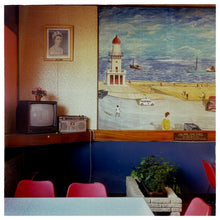 Load image into Gallery viewer, Photograph by Richard Heeps. This photo from 1986 is taken in a cafe which has a picture of Fleetwood beach erected In Memoriam which hangs next to a picture of Queen Elizabeth II.  Below is a shelf with a television and alongside that a radio.  In the room are cafe-style plastic seating and chairs.  