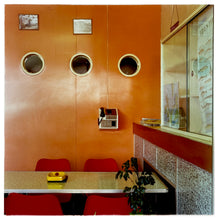Load image into Gallery viewer, Photograph by Richard Heeps.  Taken in the Fisherman&#39;s Mission cafe, on the wall are three portholes in an orange-coloured wooden wall.  On the wall is a payphone and this part of the room holds a cafe-style table and chairs, on the table is a yellow ashtray.