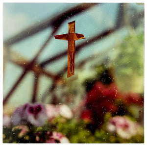 Photograph by Richard Heeps. A copper-coloured cross is suspended on frosted greenhouse glass, behind the glass there are the vague forms of the greenhouse flowers.