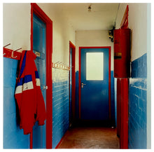 Load image into Gallery viewer, Photograph by Richard Heeps.  A hallway in a scouts hut with painted walls, the top half of the walls are painted cream and the bottom are painted blue.  There are two blue painted doors which have a red door frame.  There are red painted coat hooks on the left hand side on the wall on which is hanging a jacket made up of nearly identical red and blue colours.
