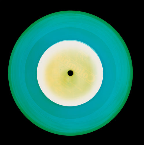 Photograph by Natasha Heidler and Richard Heeps. A Turquoise vinyl record with a yellow and white record label sits on a black background.