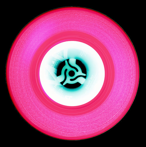 Photograph by Natasha Heidler and Richard Heeps.  A pink vinyl record with thin white grooves and a white label sits on a black background.