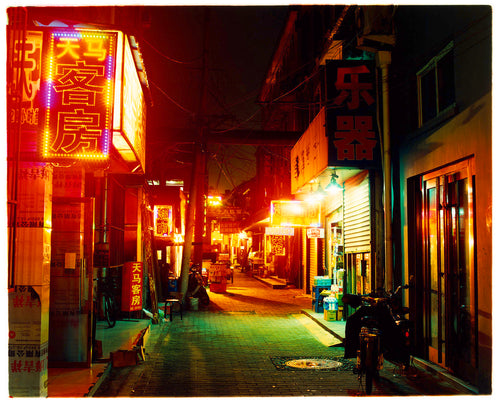 Photograph by Richard Heeps.  A narrow road in Hutong, brightly lit orange signs light the way down the road.