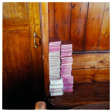 Load image into Gallery viewer, Photograph by Richard Heeps.  Stacked up in the corner of a wooden pew next to a wooden door is two columns of hymn books.  The hymn books are red hard back some with white paper edging and some with pink.