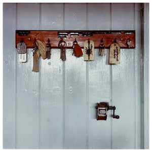 A vintage photograph of a dark wooden key holder carrying a selection of keys and fobs.  The holder is fixed on a white-painted wooden slated wall.  There is a pencil sharpener attached to the wall also, below the key holder.  Photograph by Richard Heeps.