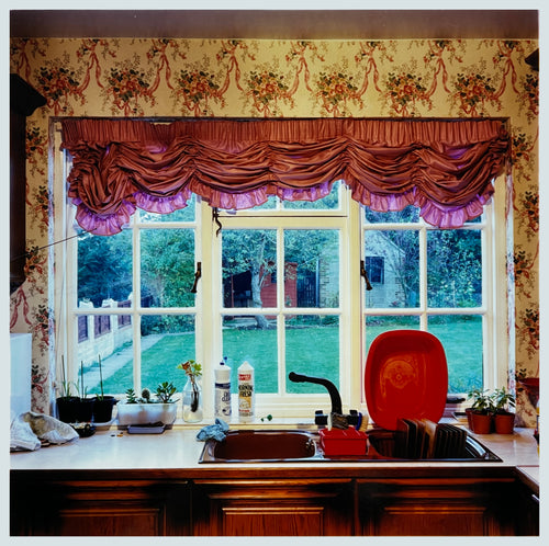 Photograph by Richard Heeps. A kitchen sink from 1989 with brown taps and bowl, and surrounded by two bottles of washing up liquid and a red washing up bowl.  The window above the sink looks out on a neat garden and is topped with orange ruched curtains.  The wallpaper has a sequence of orange flowers linked with orange ribbon.  
