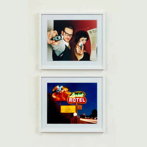 A couple look into the camera in burlesque costume against a red wall. Contemporary portrait photography by Richard Heeps framed in white