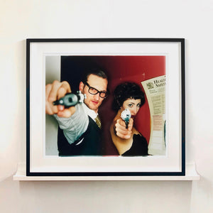 A couple look into the camera in burlesque costume against a red wall. Contemporary portrait photography by Richard Heeps framed in black