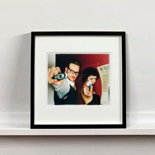 Load image into Gallery viewer, A couple look into the camera in burlesque costume against a red wall. Contemporary portrait photography by Richard Heeps framed in black