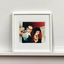 Load image into Gallery viewer, A couple look into the camera in burlesque costume against a red wall. Contemporary portrait photography by Richard Heeps framed in white