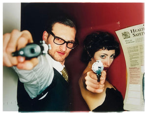 A couple look into the camera in burlesque costume against a red wall. Contemporary portrait photography by Richard Heeps