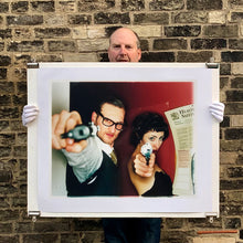 Load image into Gallery viewer, A couple look into the camera in burlesque costume against a red wall. Contemporary portrait photography by Richard Heeps