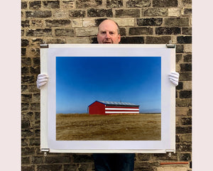 'Stars & Stripes Barn' shows a depiction of the American flag painted on this isolated building in Oakhurst, California. It sits on the horizon against a vast bright blue sky. This artwork is part of Richard Heeps' 'Dream in Colour' series.