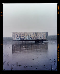 Photograph by Richard Heeps.  This photograph is tones of grey.  DEJAVU is painted on the side of a lorry trailer which sits in a grey watery fen, the sky is grey in the background. This photograph includes a film rebate.