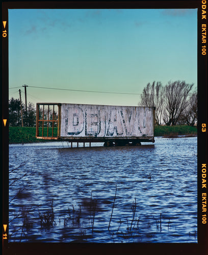 Photograph by Richard Heeps. A broken trailer sits in blue water, the side of the trailer has the faded wording DEJAVU.