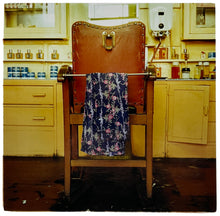 Load image into Gallery viewer, Photograph by Richard Heeps. An antique barbers chair, with a customer robe draped over the front, sits in front of an aged yellow side board on which sits many barber type lotions. There is a water heater sitting on the wall above the sideboard