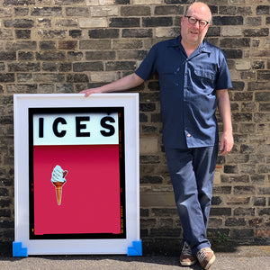 Photograph by Richard Heeps.  Richard Heeps holds a white framed print. At the top of the print, black letters spell out ICES and below is depicted a 99 icecream cone sitting left of centre against a raspberry coloured background.  