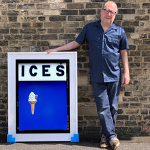 Load image into Gallery viewer, Photograph by Richard Heeps.  Richard Heeps holds a white framed print. At the top of the print, black letters spell out ICES and below is depicted a 99 icecream cone sitting left of centre against a blue coloured background.  