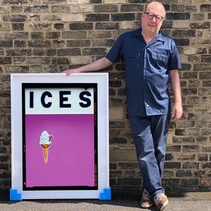 Photograph by Richard Heeps.  Richard Heeps holds a white framed print. At the top of the print, black letters spell out ICES and below is depicted a 99 icecream cone sitting left of centre against a plum coloured background.  