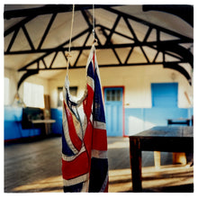 Load image into Gallery viewer, Photograph by Richard Heeps.  A Union Jack hangs from the rafters of a Scout Hut.  The hut has blue doors, a wooden floor, an old wooden table and wooden rafters. 