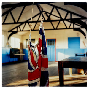 Photograph by Richard Heeps.  A Union Jack hangs from the rafters of a Scout Hut.  The hut has blue doors, a wooden floor, an old wooden table and wooden rafters. 