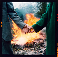 Load image into Gallery viewer, Two people in in winter coats hold hand by a fire outdoors at Wysing Arts Centre Cambridge.