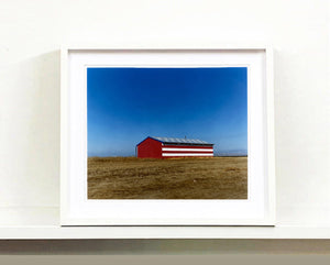 'Stars & Stripes Barn' shows a depiction of the American flag painted on this isolated building in Oakhurst, California. It sits on the horizon against a vast bright blue sky. This artwork is part of Richard Heeps' 'Dream in Colour' series.